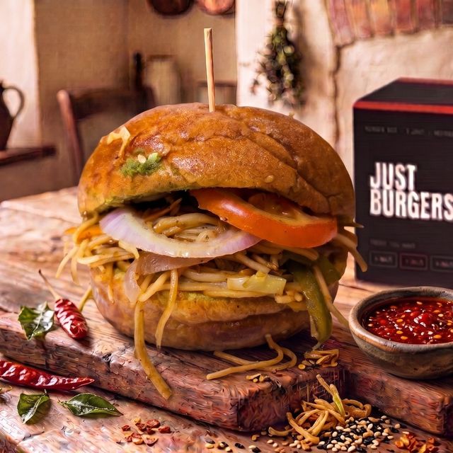 The No.4 Chowmein burger on a wooden board — a toasted bun stuffed with wok-tossed noodles, capsicum, tomato and onion, red chilli sauce beside it.